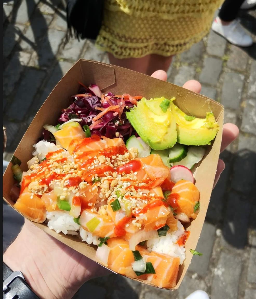 Handheld cardboard container of salmon poke with avocado, rice, red cabbage, and spicy sauce.