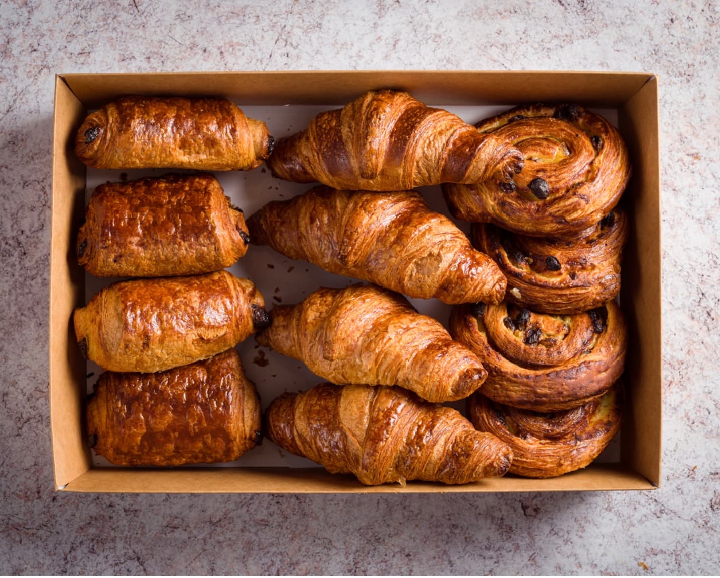 Cardboard box filled with rows of fresh croissants, pain au chocolat, and pain aux raisins.