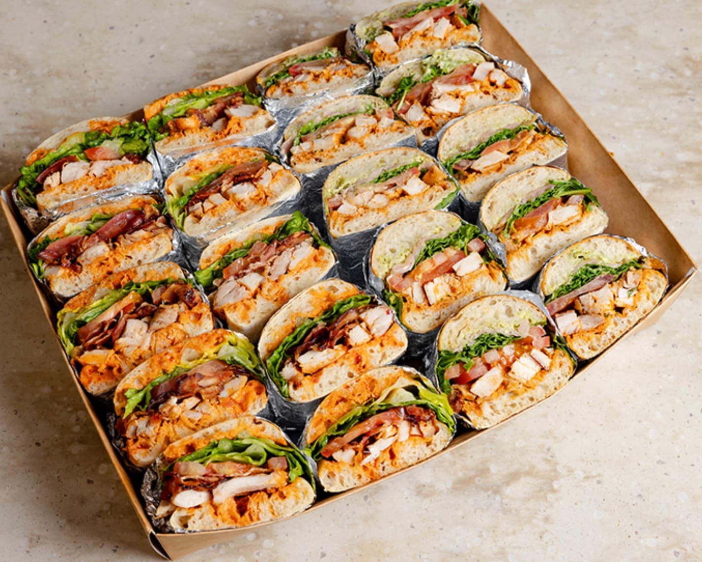 Cardboard catering box filled with rows of chicken, bacon, lettuce, and tomato sandwich halves.