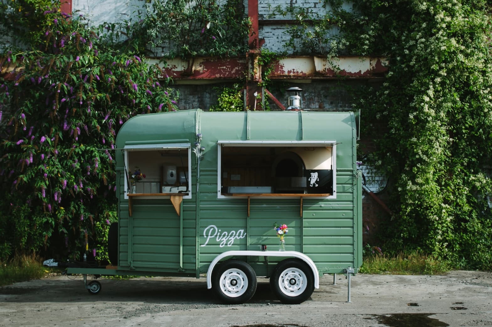 Sage green mobile pizza trailer with open serving windows parked against a lush, vine-covered wall.