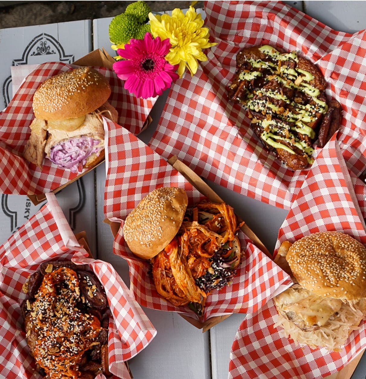 Five trays of burgers and loaded sides on red checkered paper with colorful flowers.