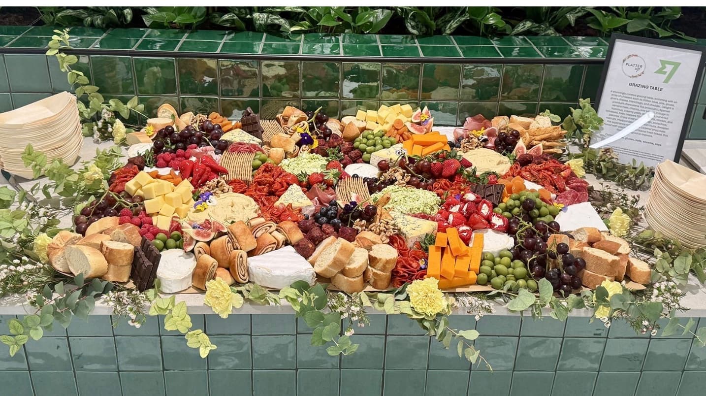 Abundant grazing table with cheeses, meats, fruits, and crackers on a green tiled counter.