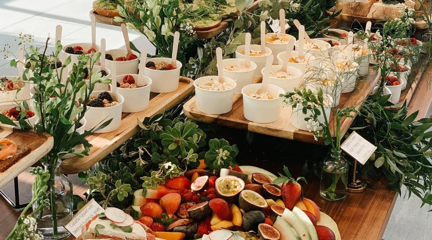 Breakfast buffet featuring yogurt cups, fresh fruit platters, and greenery on wooden serving boards.